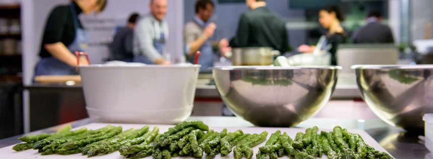 front view of food on a cooking board, with students learning how to cook in the background. Italy